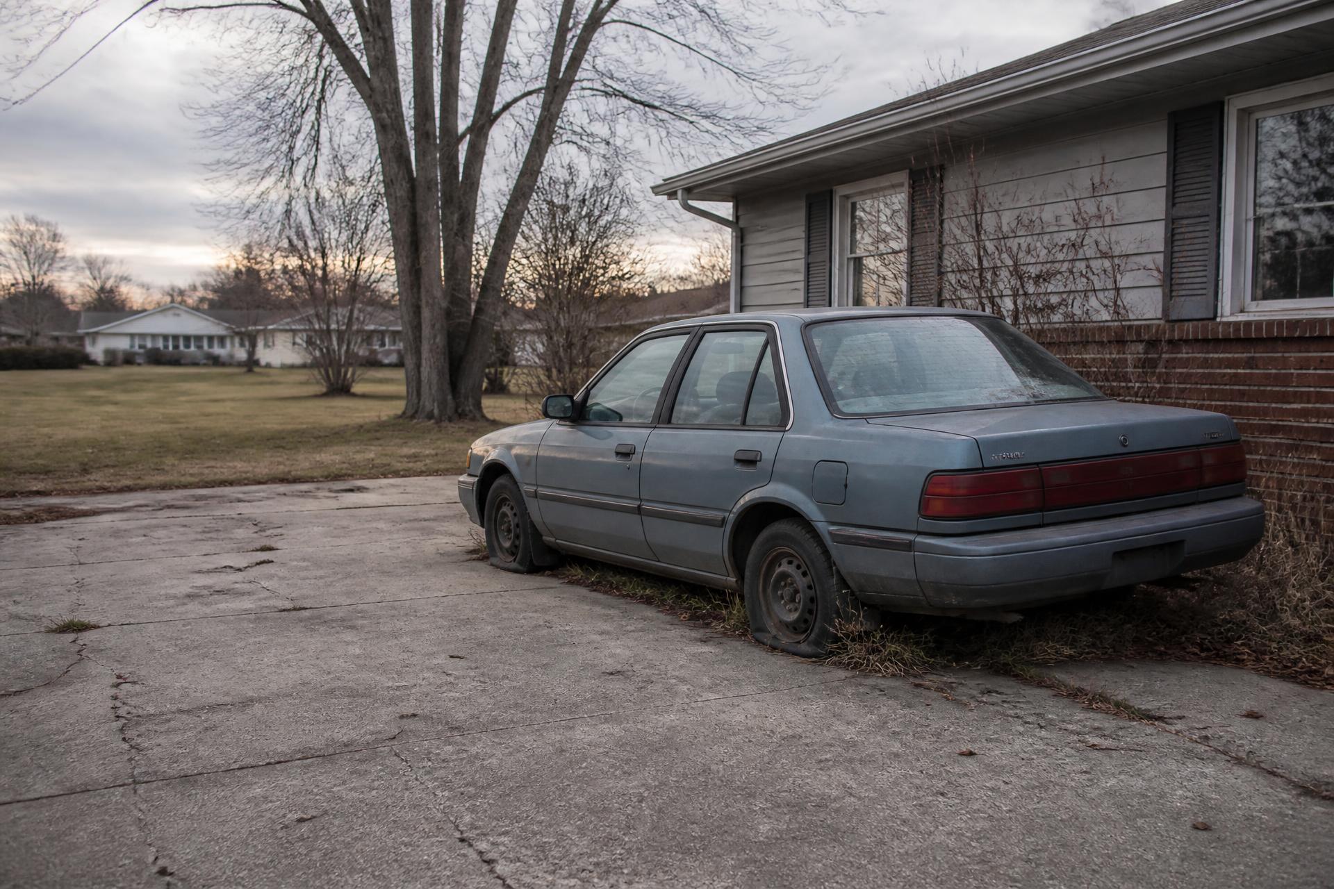 An older sedan with flat tires sitting unused in a residential driveway